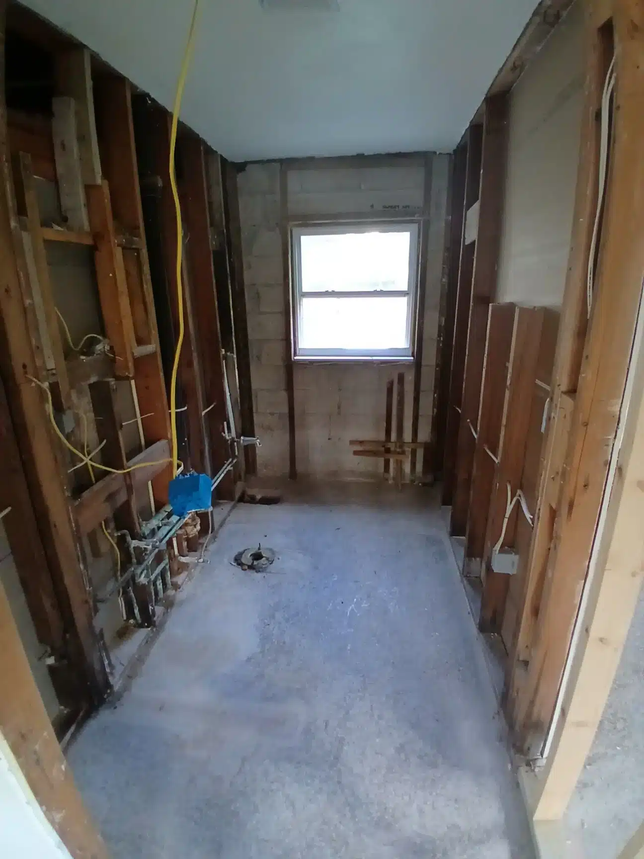 A narrow hallway or corridor under renovation shows exposed wooden door frames and studs lining both walls, with yellow electrical wiring visible on the left side. The concrete subfloor is bare, and a window at the end of the hallway provides natural light, while a blue bucket and construction materials are scattered throughout the space. The ceiling is unfinished with visible framing and the walls appear to be in mid-stage demolition or reconstruction.