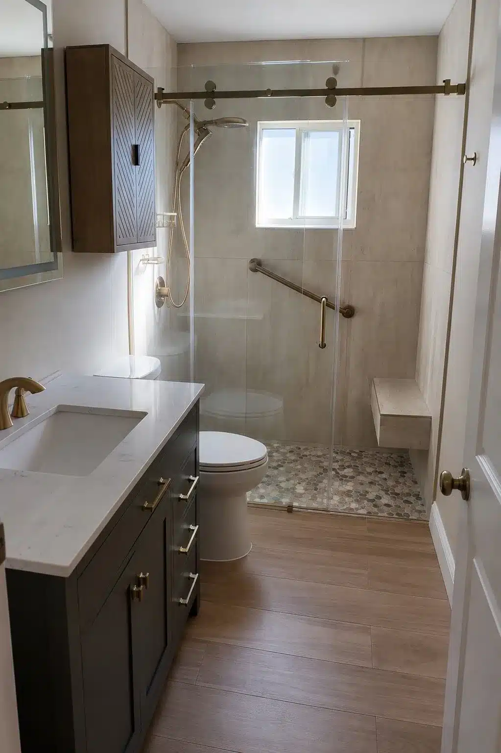 Dark vanity beside a toilet and glass-enclosed shower with beige tile, pebble floor, grab bar and bench.