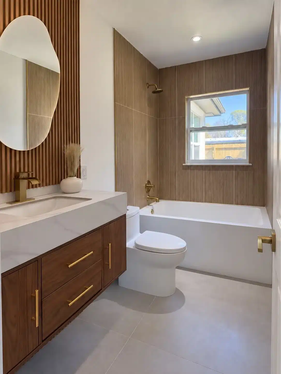 Wood vanity with white countertop, oval mirror, brass fixtures and a white tub beneath a window.