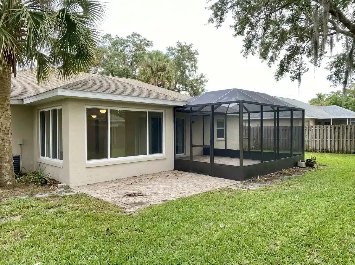 Cream-colored house with black-framed screened porch and paver patio surrounded by grass and trees.
